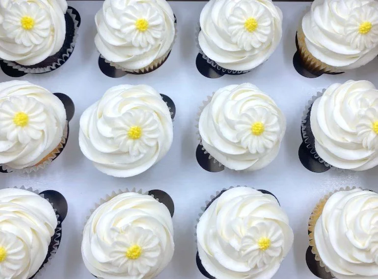 Multiple cupcakes with white frosting and small yellow flower decorations on top.