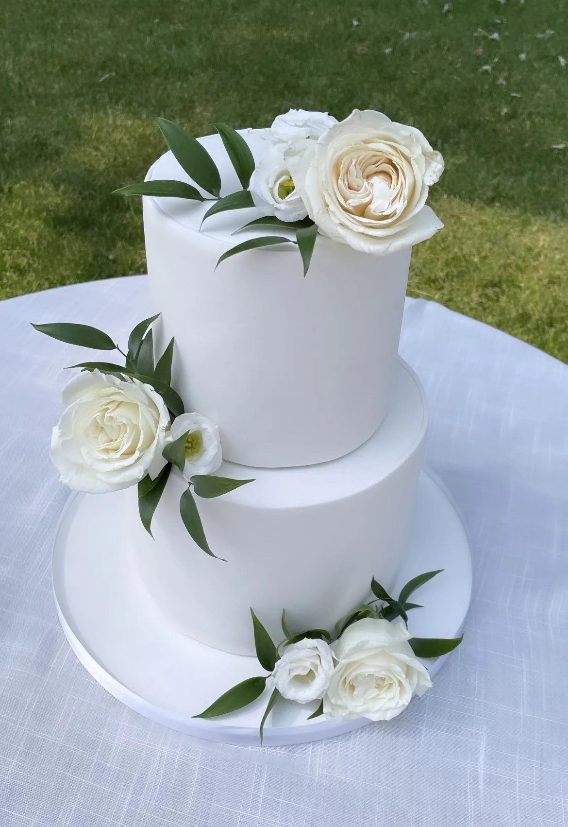 Two-tier white wedding cake decorated with white roses and green leaves, set outdoors on a white tablecloth.