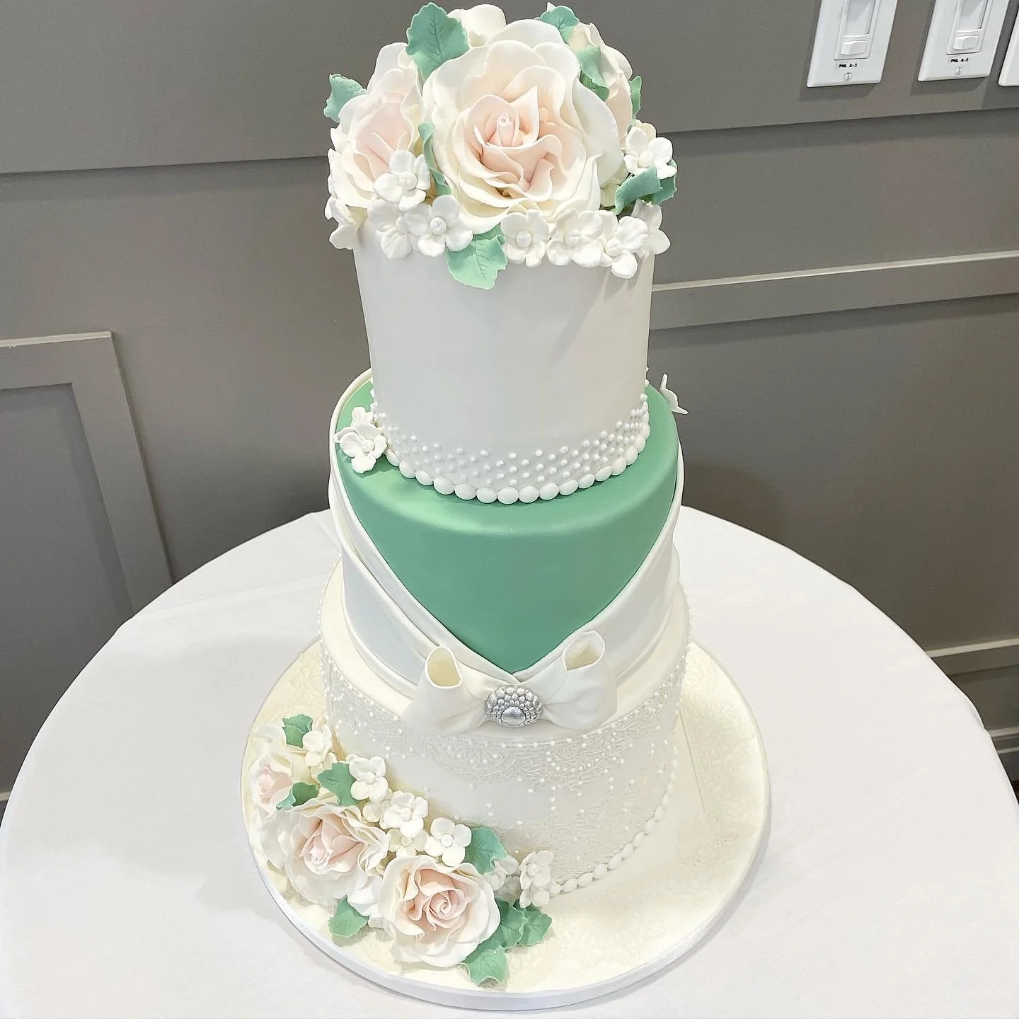 A three-tiered wedding cake decorated with pink roses, white flowers, green leaves, a white bow, and pearl-like accents.