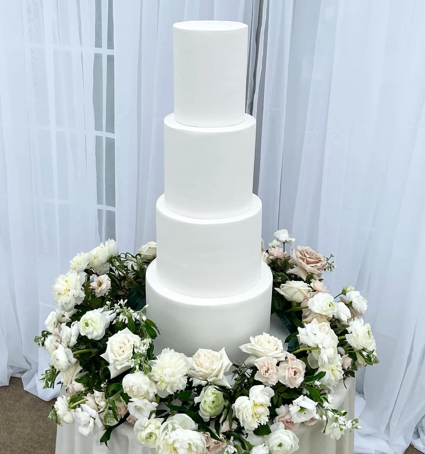 White four-tiered wedding cake surrounded by a wreath of white and pale pink roses and flowers on a table with white drapes in the background.