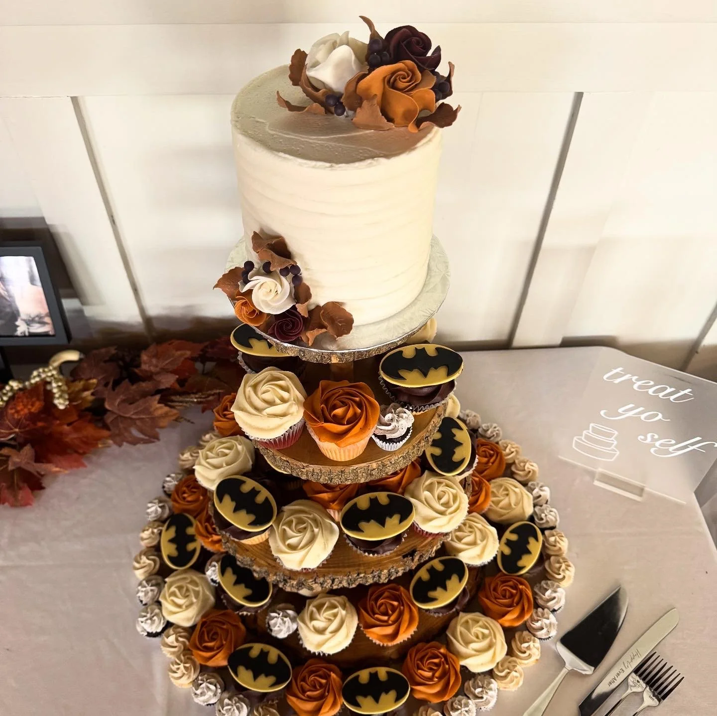 A multi-tiered cake decorated with Batman-themed cupcakes, chocolate roses, and fall leaves.