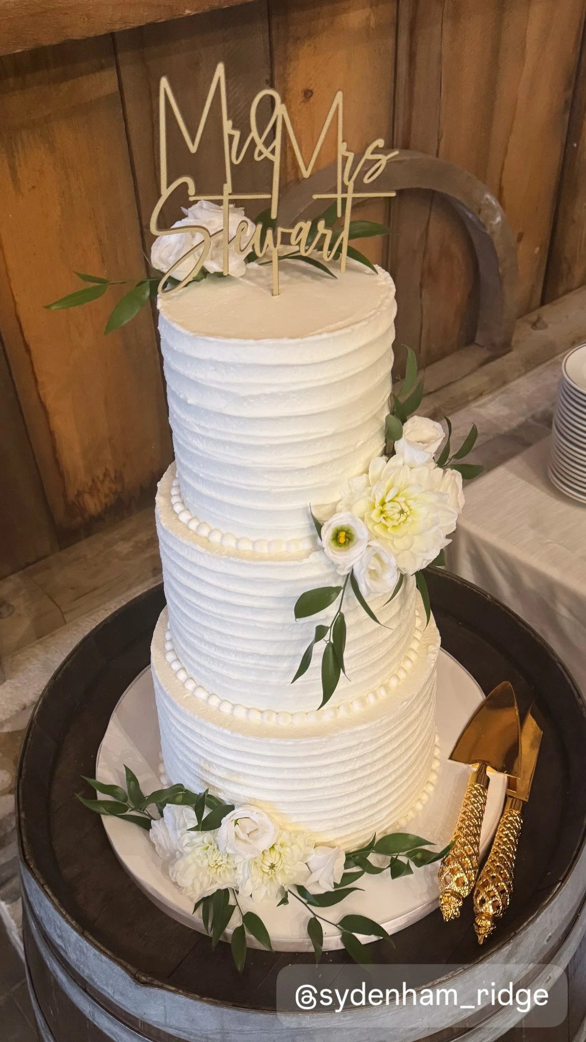 A three-tiered white wedding cake decorated with white flowers and greenery, topped with a cake topper.