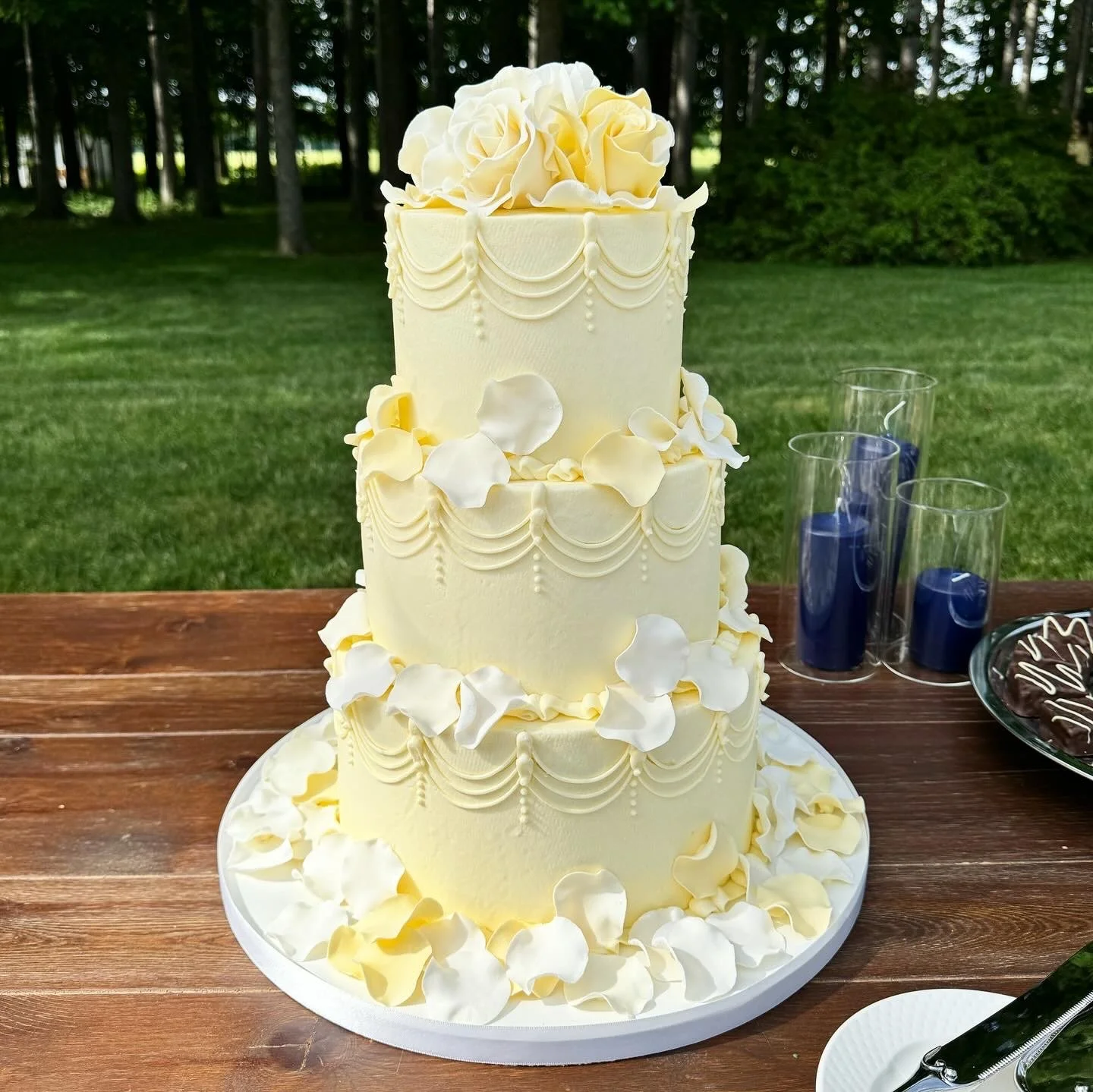 Three-tier white wedding cake decorated with flower petals and a large floral topper, set on a wooden table outdoors with grass and trees in the background.
