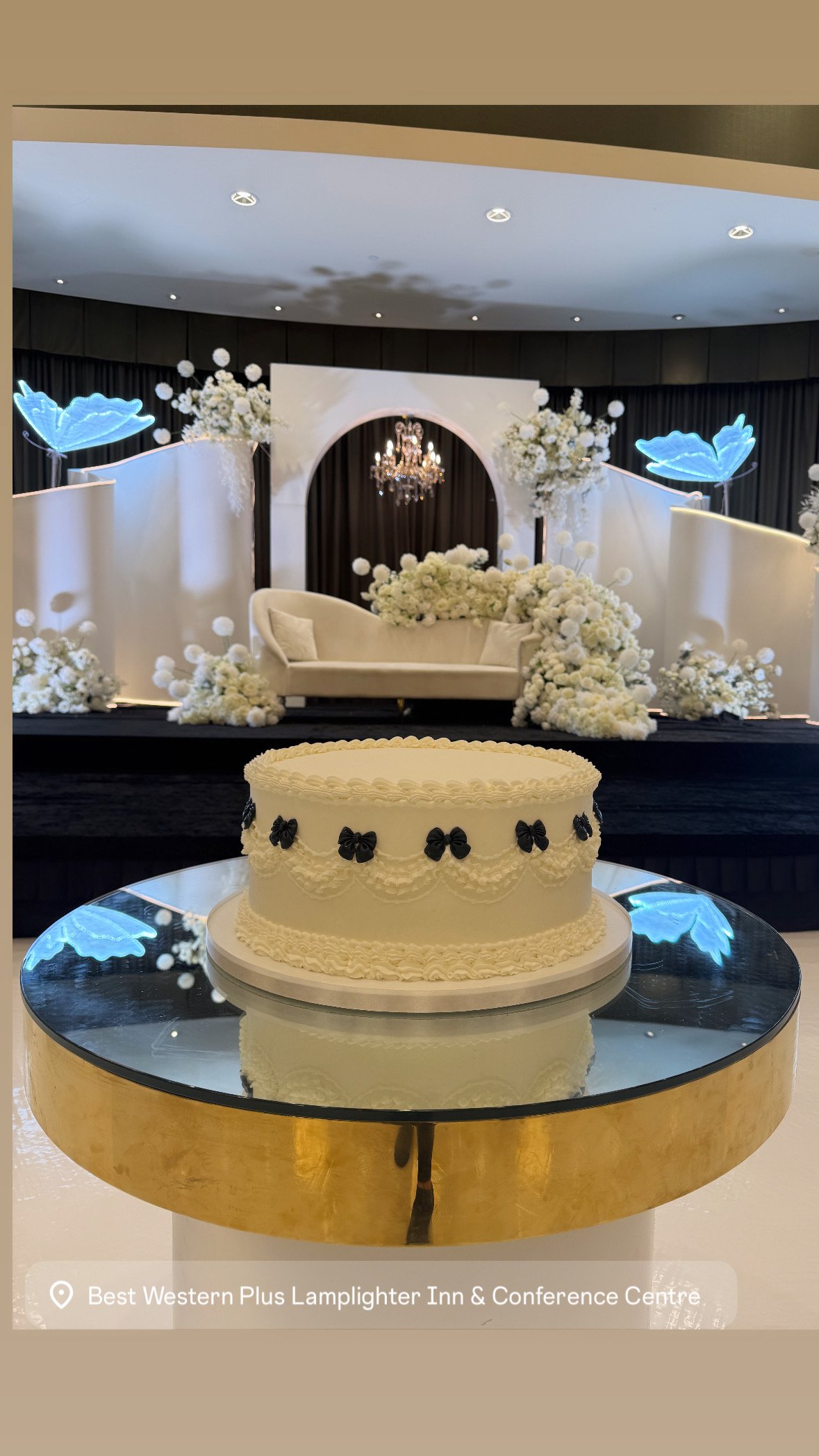 A wedding cake with black bows on a reflective glass table, with a wedding stage decorated with white flowers, a white sofa, and neon blue decorations in the background at the Best Western Plus Lamplighter Inn & Conference Centre.