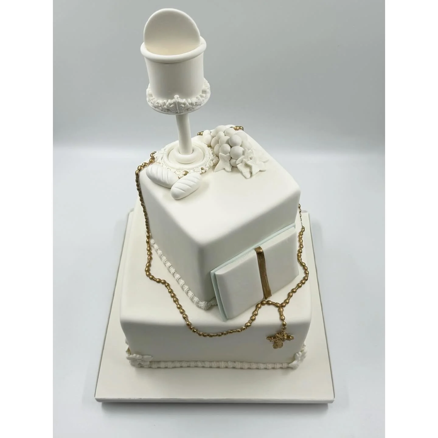 White two-tiered cake decorated with religious symbols including a chalice, a book, and a rosary.