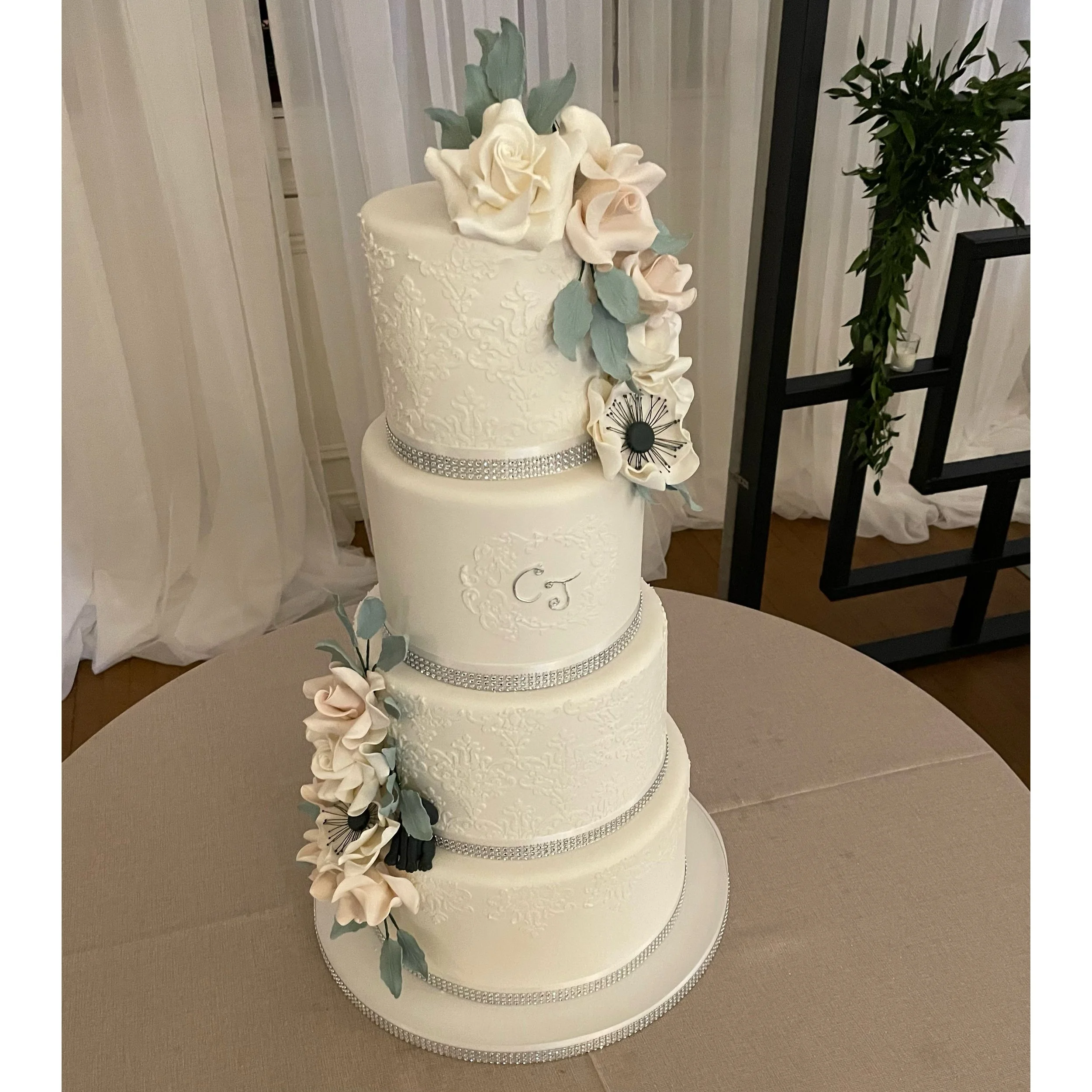 A tall, four-tiered white wedding cake decorated with intricate piping, silver ribbon accents, and cascading sugar flowers in soft pastel colors.