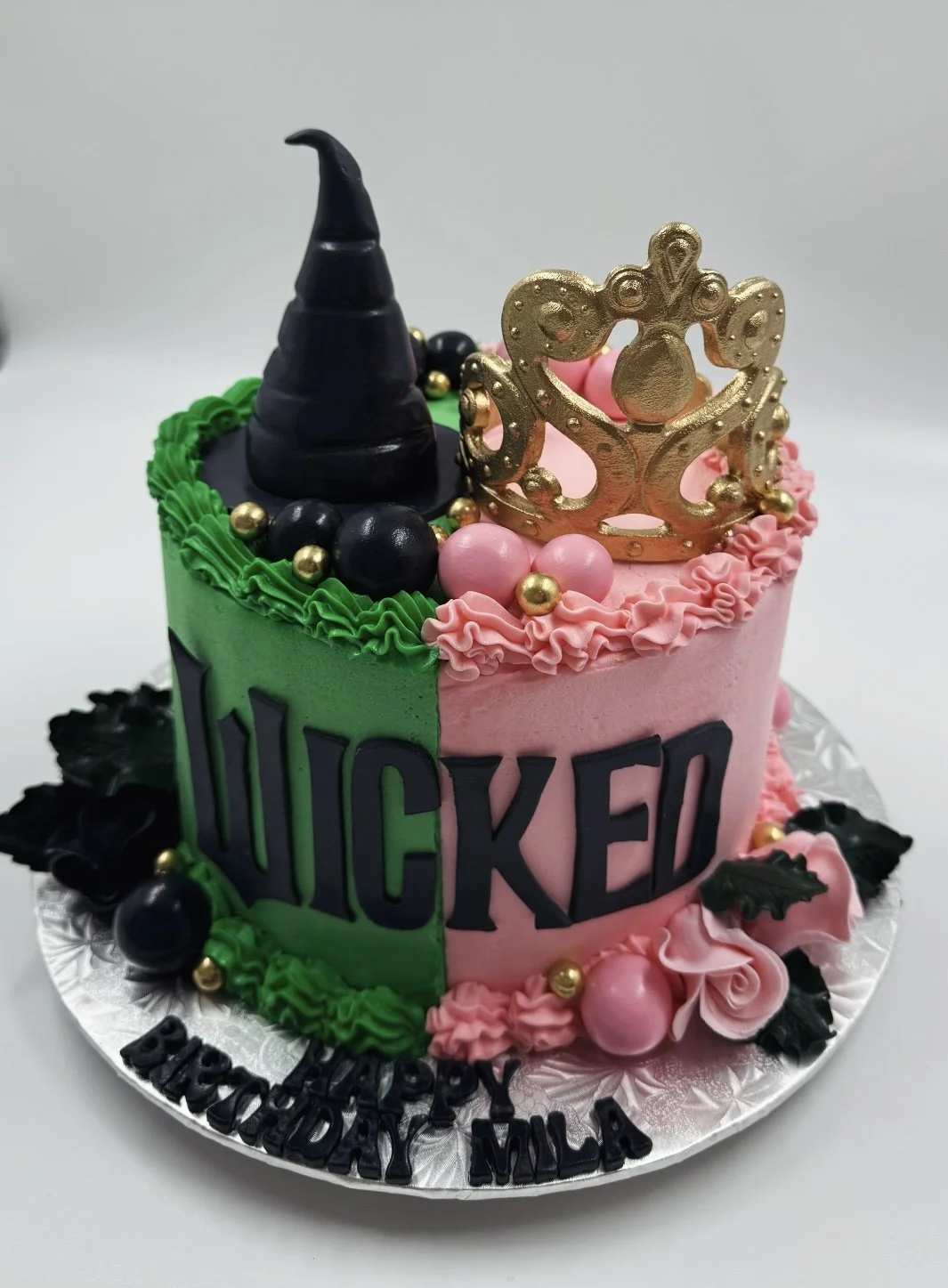 A colorful birthday cake decorated with a black witch's hat, a gold crown, pink and black icing, and various jewelry-style decorations, with black lettering that spells out "WICKED".