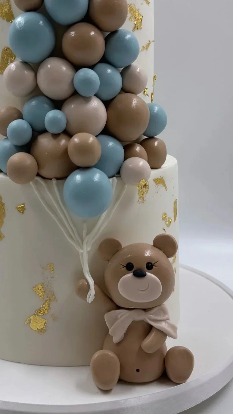 Cake decorated with pastel-colored spheres resembling balloons, a cute teddy bear figure with a bow, and gold leaf accents.