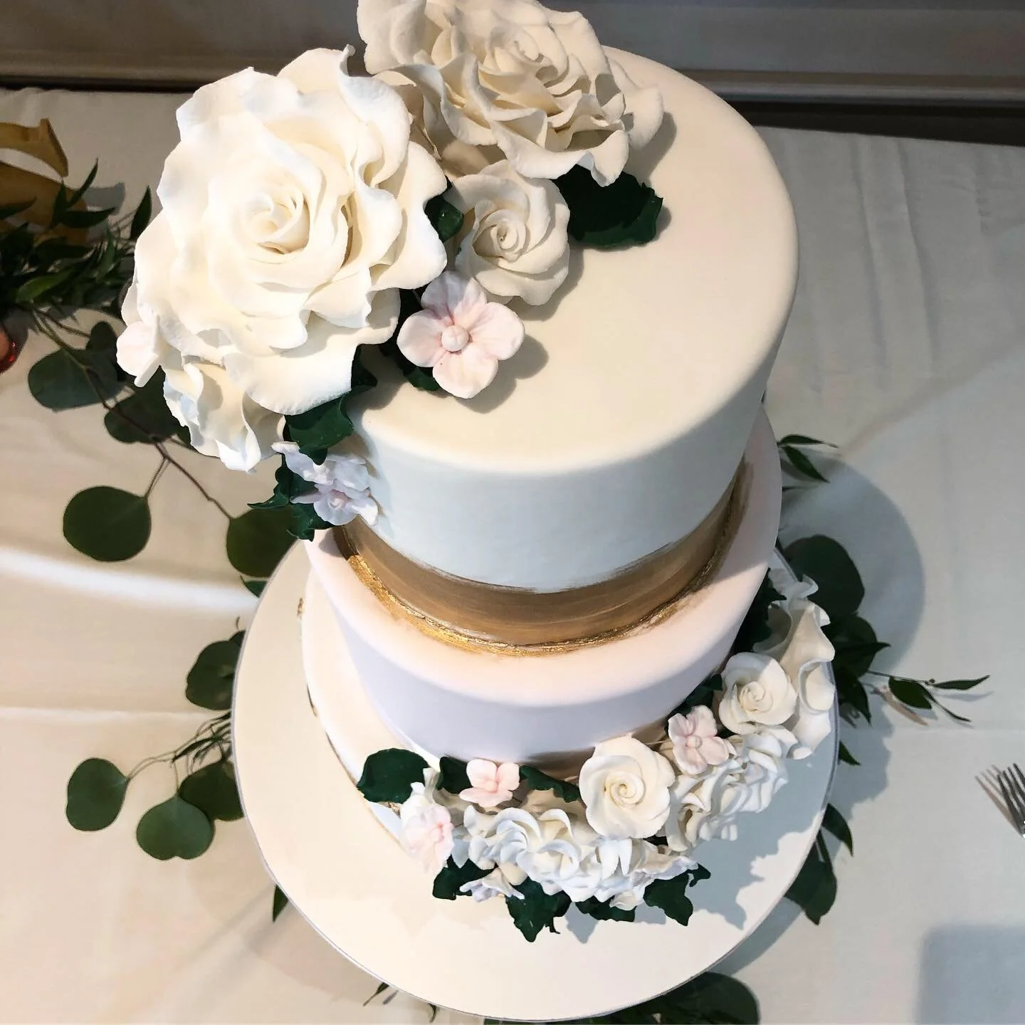 Two-tier wedding cake decorated with white and pink flowers and green leaves, with a gold band around the middle layer.