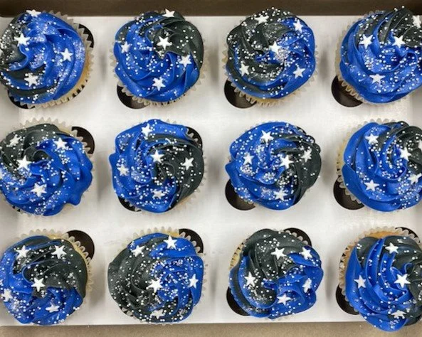  twelve cupcakes decorated with blue and black swirled frosting, topped with white star and dot sprinkles, arranged on a white surface.