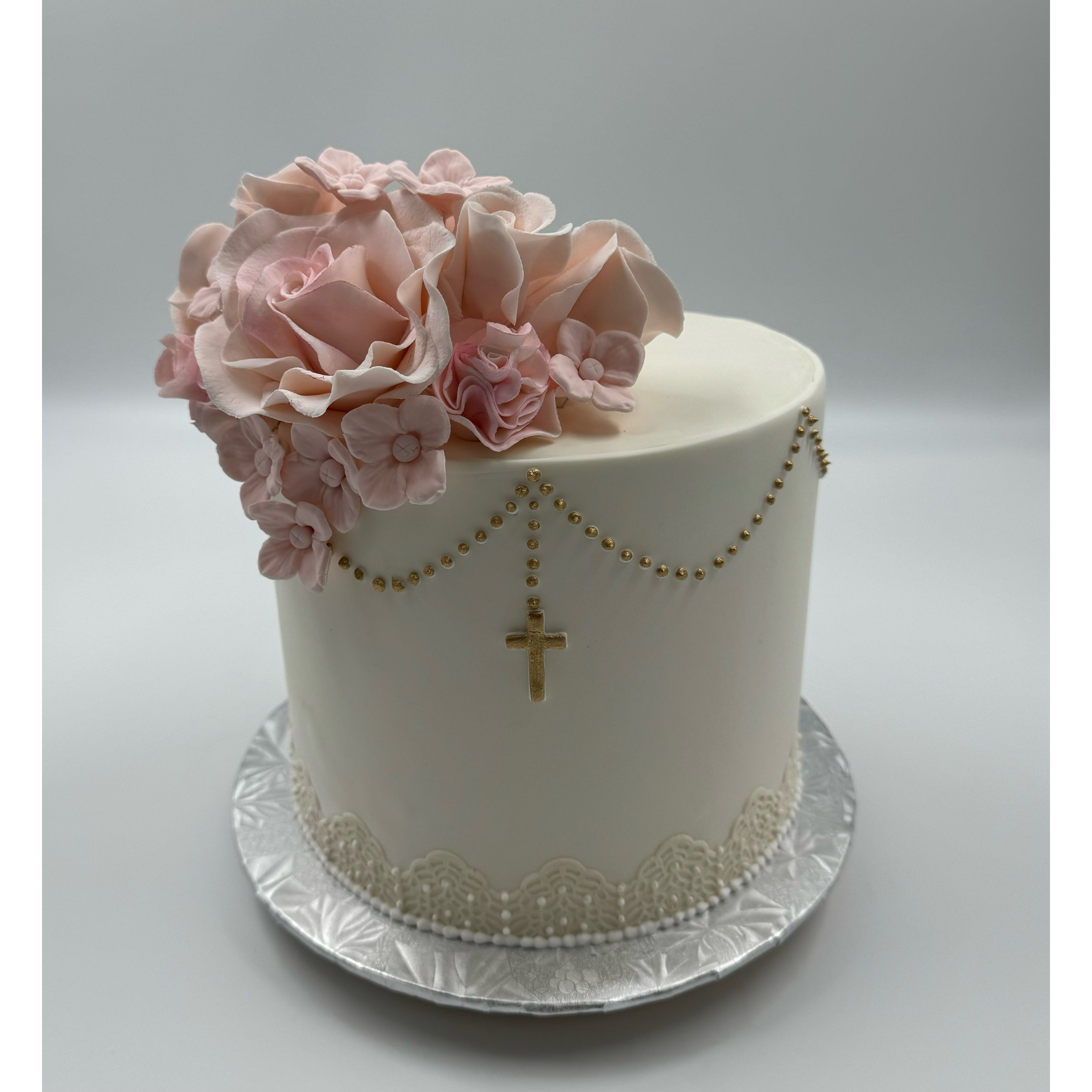 A white cake decorated with pink sugar flowers and gold accents, including a gold cross pendant and a gold beaded chain. The cake sits on a silver, patterned cake board.