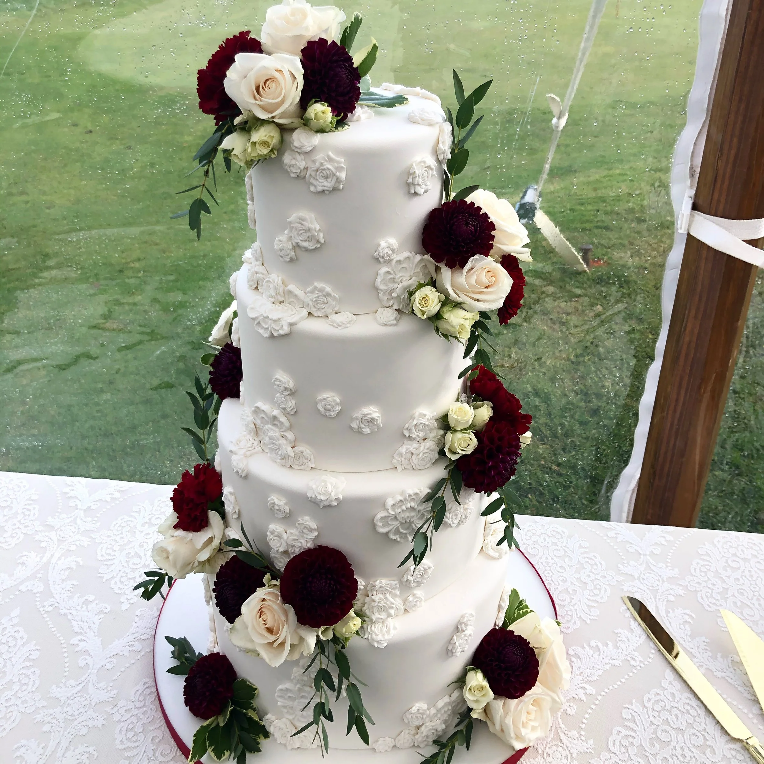 A tall, multi-tiered white wedding cake decorated with white and dark red flowers and green leaves.