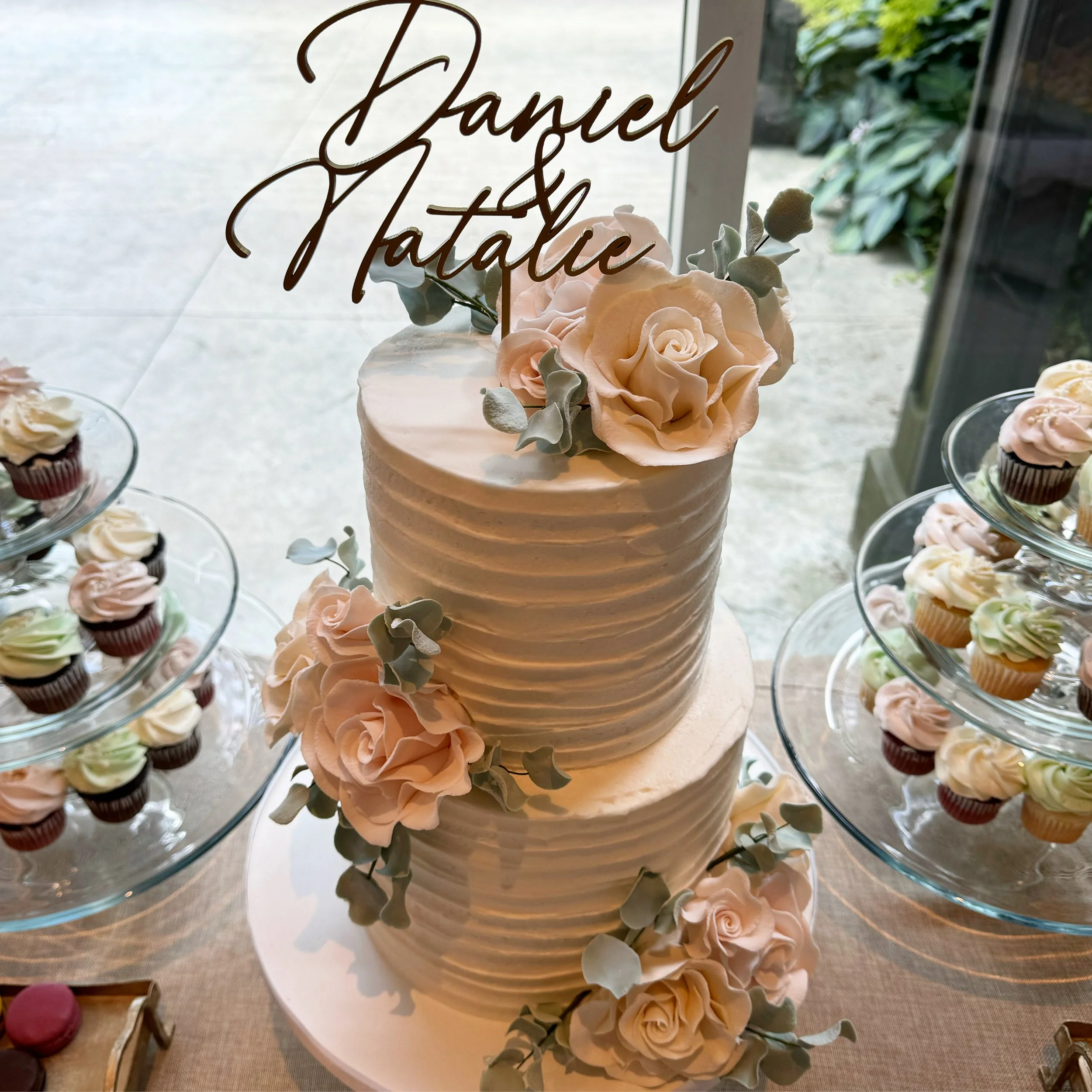 A three-tiered white wedding or celebration cake with pink roses and green foliage decorations, positioned next to glass display stands filled with assorted cupcakes.