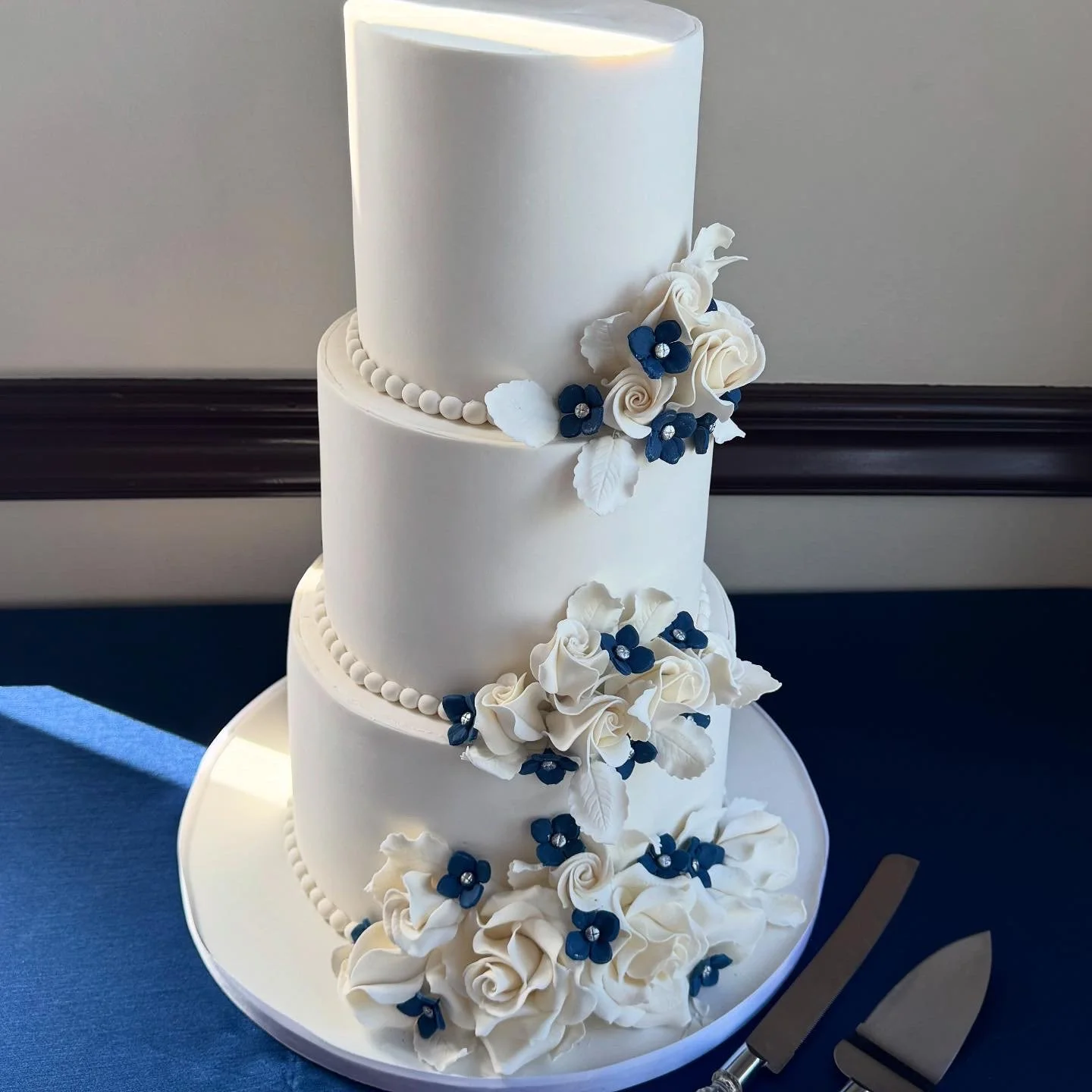 Three-tier white wedding cake decorated with white roses, dark blue flowers, and white leaves.