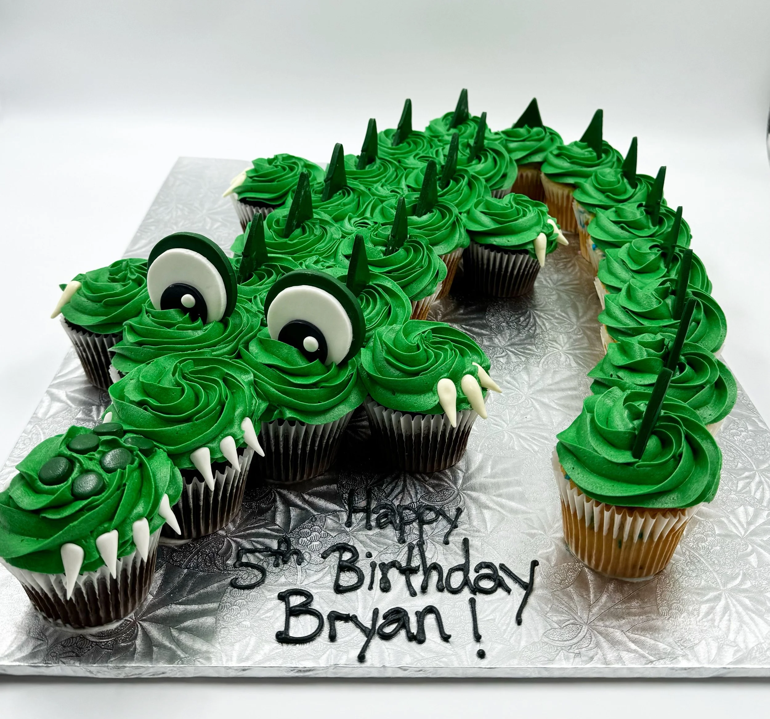 Cupcake cake decorated as a crocodile with green frosting, white and black eyes, white teeth, and green triangular spikes on its back.