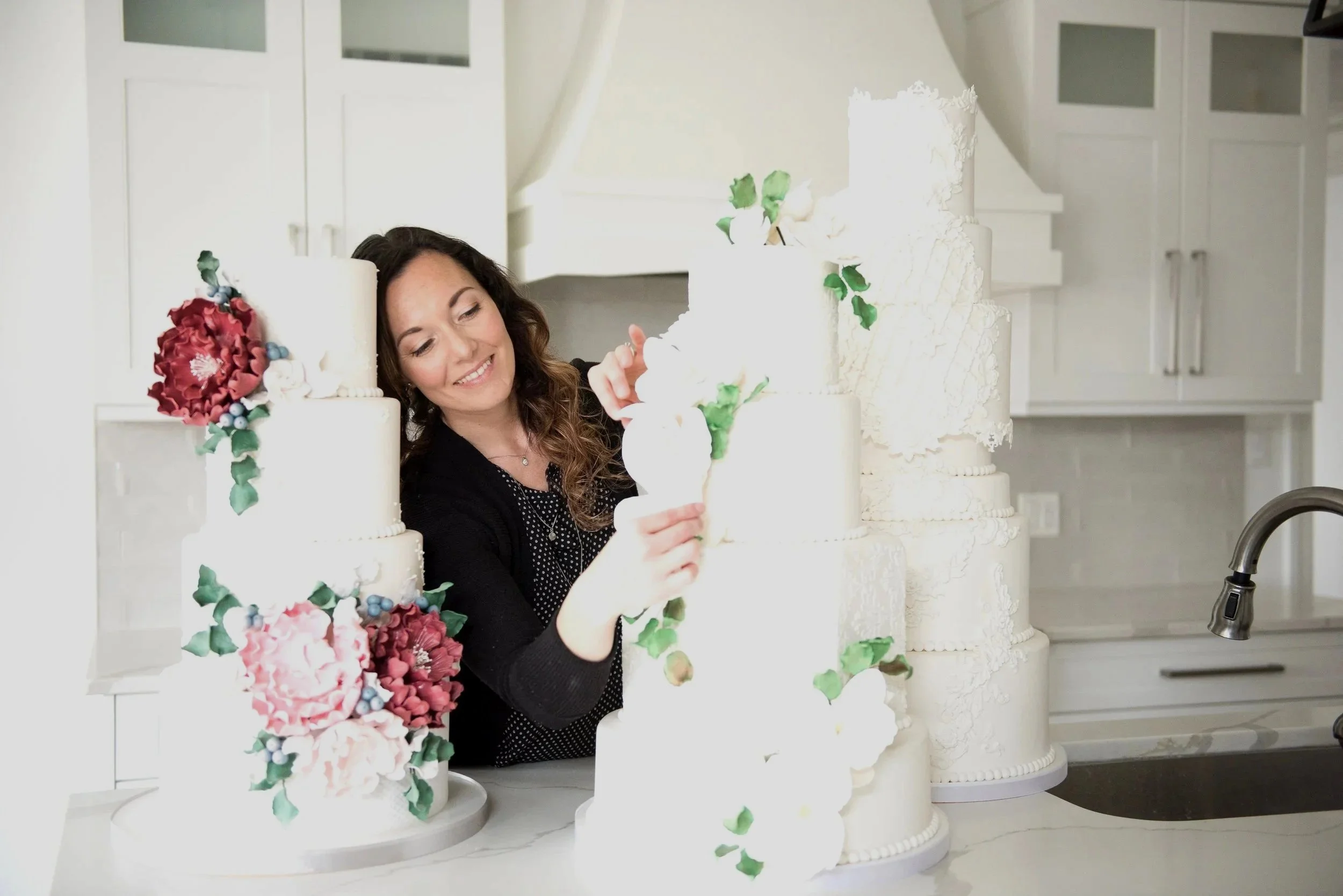 A woman decorating a large, multi-tiered white wedding cake with colorful flower decorations in a modern kitchen.