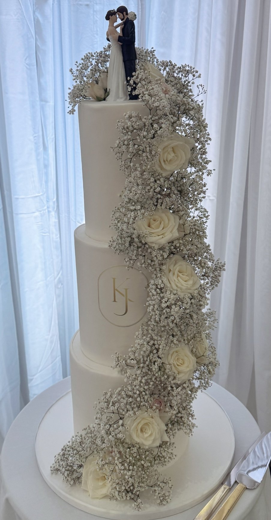A tall white wedding cake decorated with white roses and baby's breath flowers, topped with a figurine of a bride and groom, with an initial 'KJ' on the middle tier, set against a light curtain background.