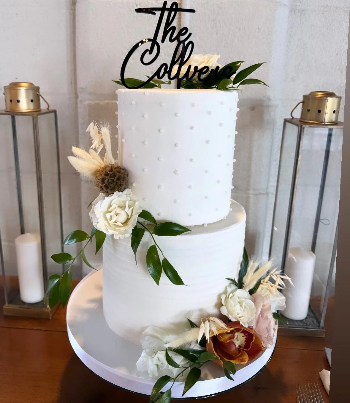 A two-tier white wedding cake decorated with white roses, greenery, and dried flowers, topped with a black cake topper.
