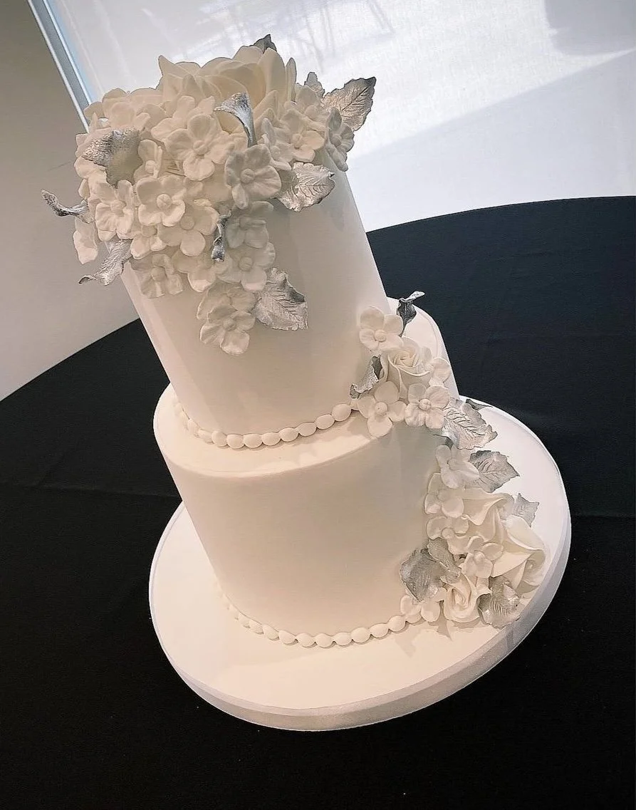 Two-tier white wedding cake decorated with white flowers and silver leaves, placed on a white cake board.