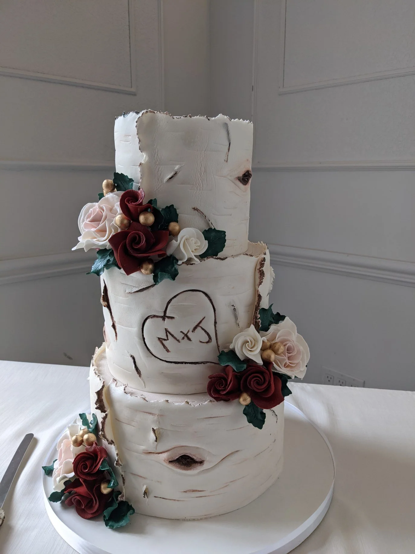Three-tier wedding cake with white textured icing resembling birch bark, decorated with red, white, and pink roses, green leaves, and gold berries.