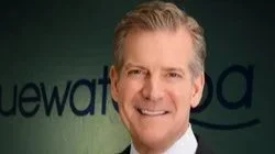 A man with gray hair smiling in front of a green background with partially visible text.