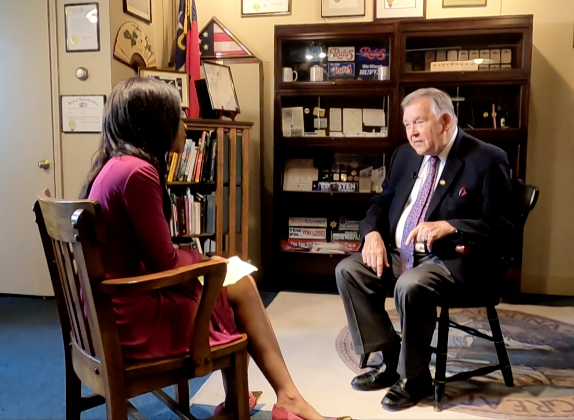 An interview setting with a female interviewer and an elderly male interviewee in an office, with books, awards, and flags in the background.