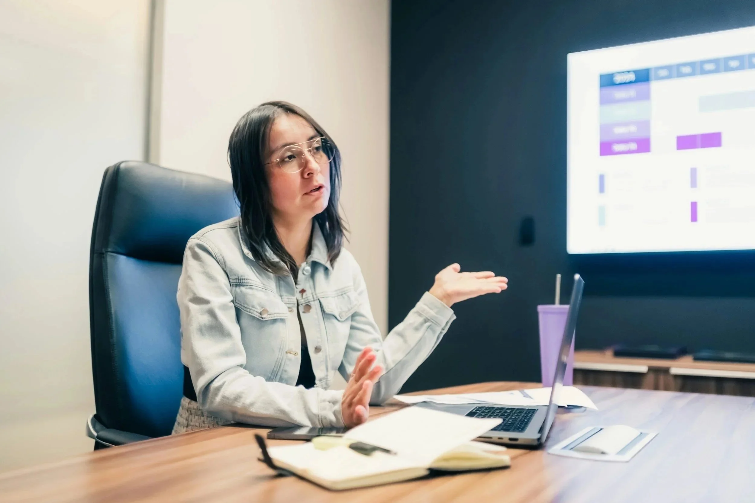 A woman with glasses and shoulder-length dark hair, wearing a light-colored jacket, sitting at a table during a meeting or presentation. She is gesturing with her hands, with an open laptop, notebooks, and a purple cup on the table. A large screen displays a colorful chart or schedule in the background.