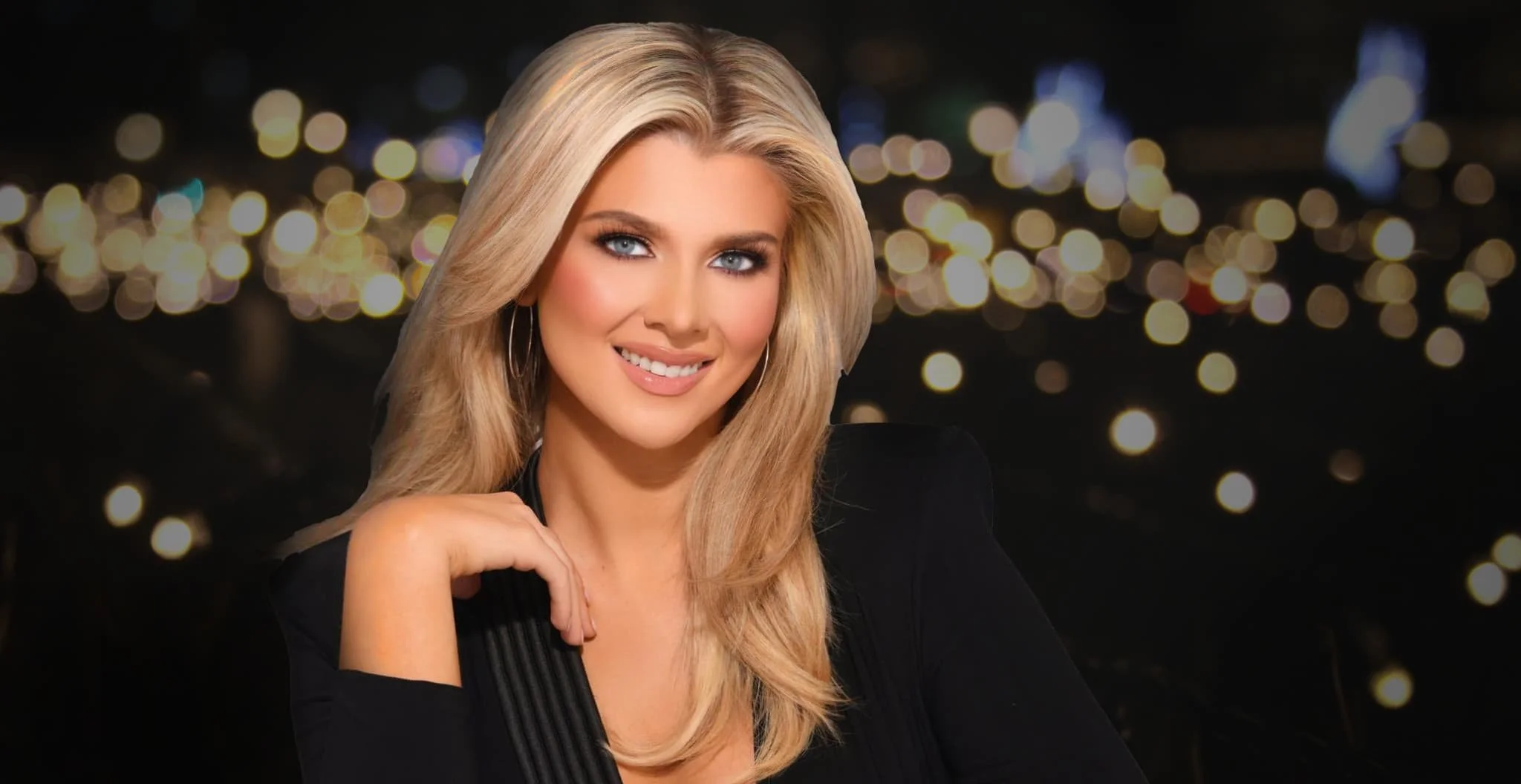 A smiling blonde woman with blue eyes and hoop earrings, wearing a black top, with a bokeh city lights background at night.