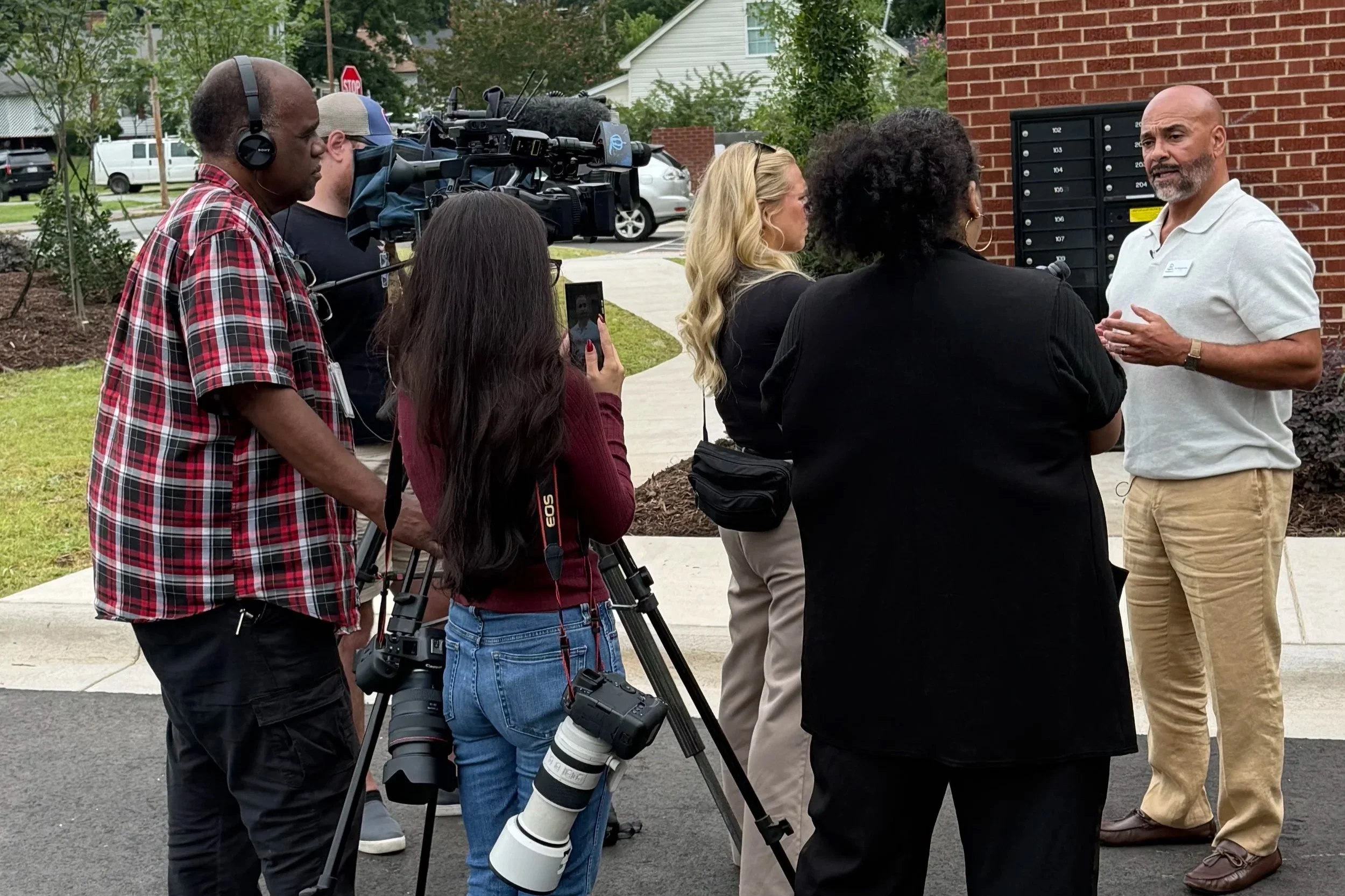 A group of people, including a cameraman, are interviewing a man outside near a brick building and mailboxs.