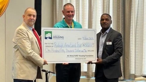 Three men, two in suits and one in a teal shirt, holding a large check for a donation from NC REALTORS' housing foundation at an indoor event.