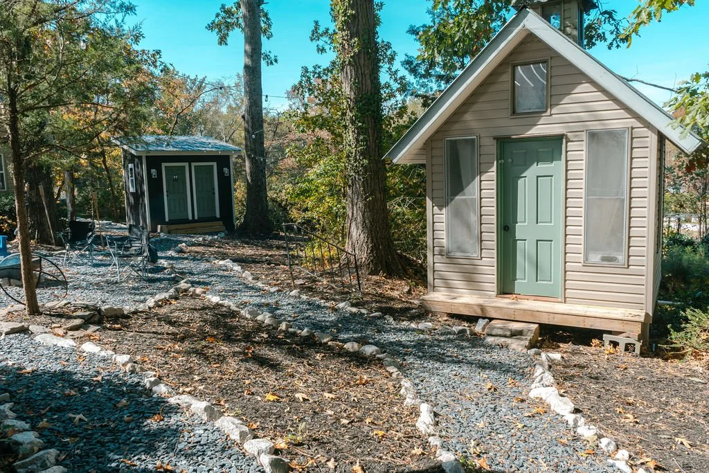 Two small houses, one in the foreground with beige siding and a green door, and a second in the background with dark siding, are situated in a wooded outdoor area. The ground is covered with gravel and stones, with a narrow pathway lined with rocks l