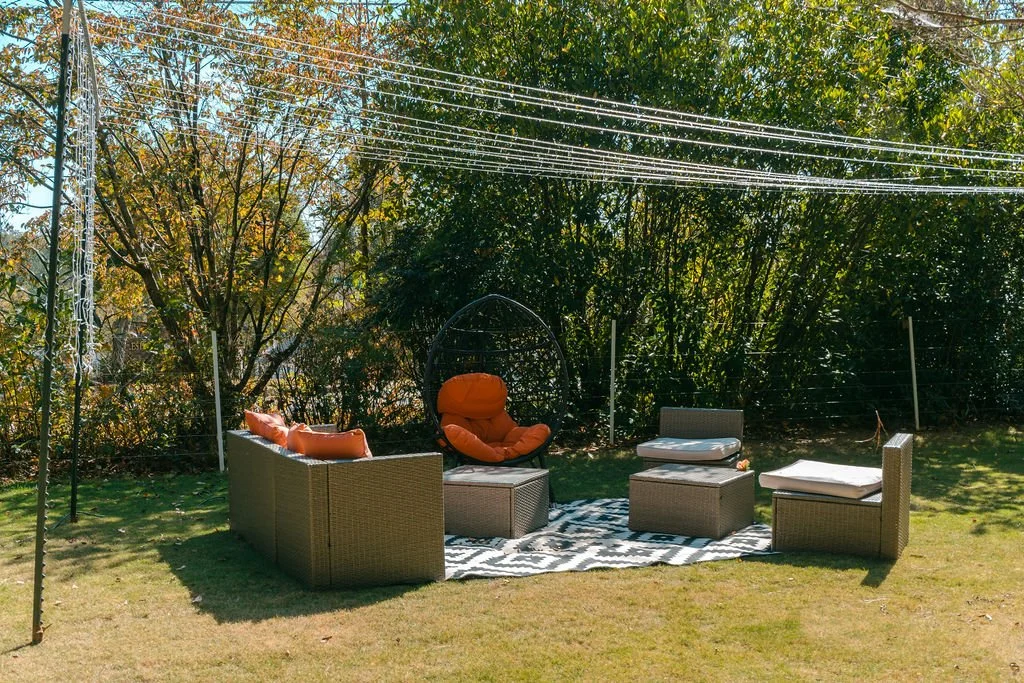Outdoor patio with wicker furniture, cushions, a black hanging chair with orange cushions, a patterned rug, and surrounded by trees and a wire fence.