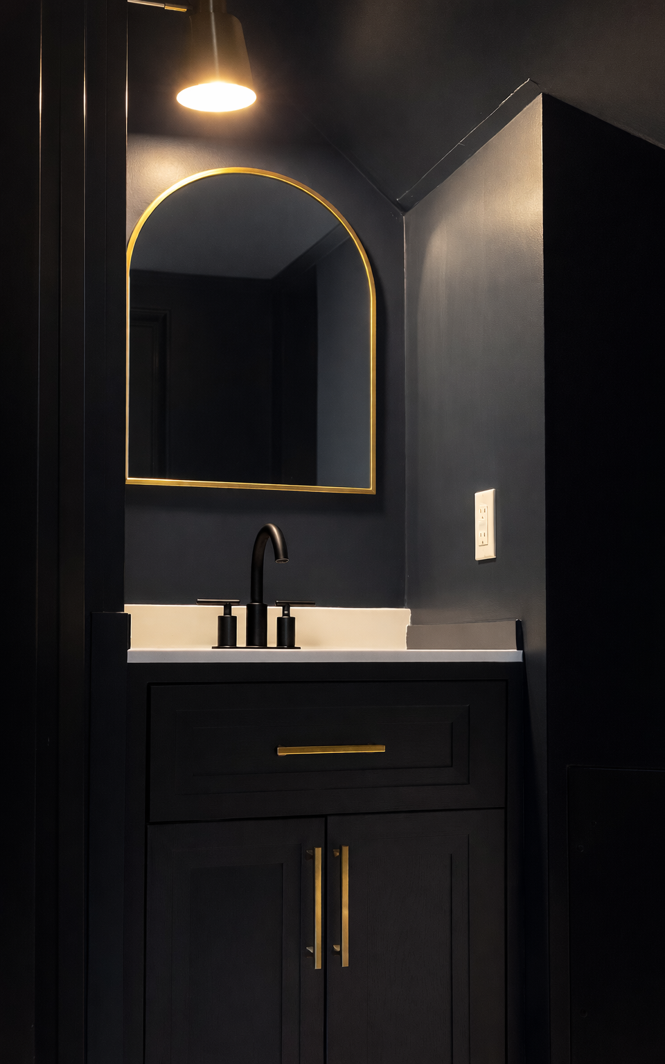 Dark bathroom vanity with black cabinet, white countertop, gold handle, black faucet, rectangular mirror with gold trim, wall-mounted light above