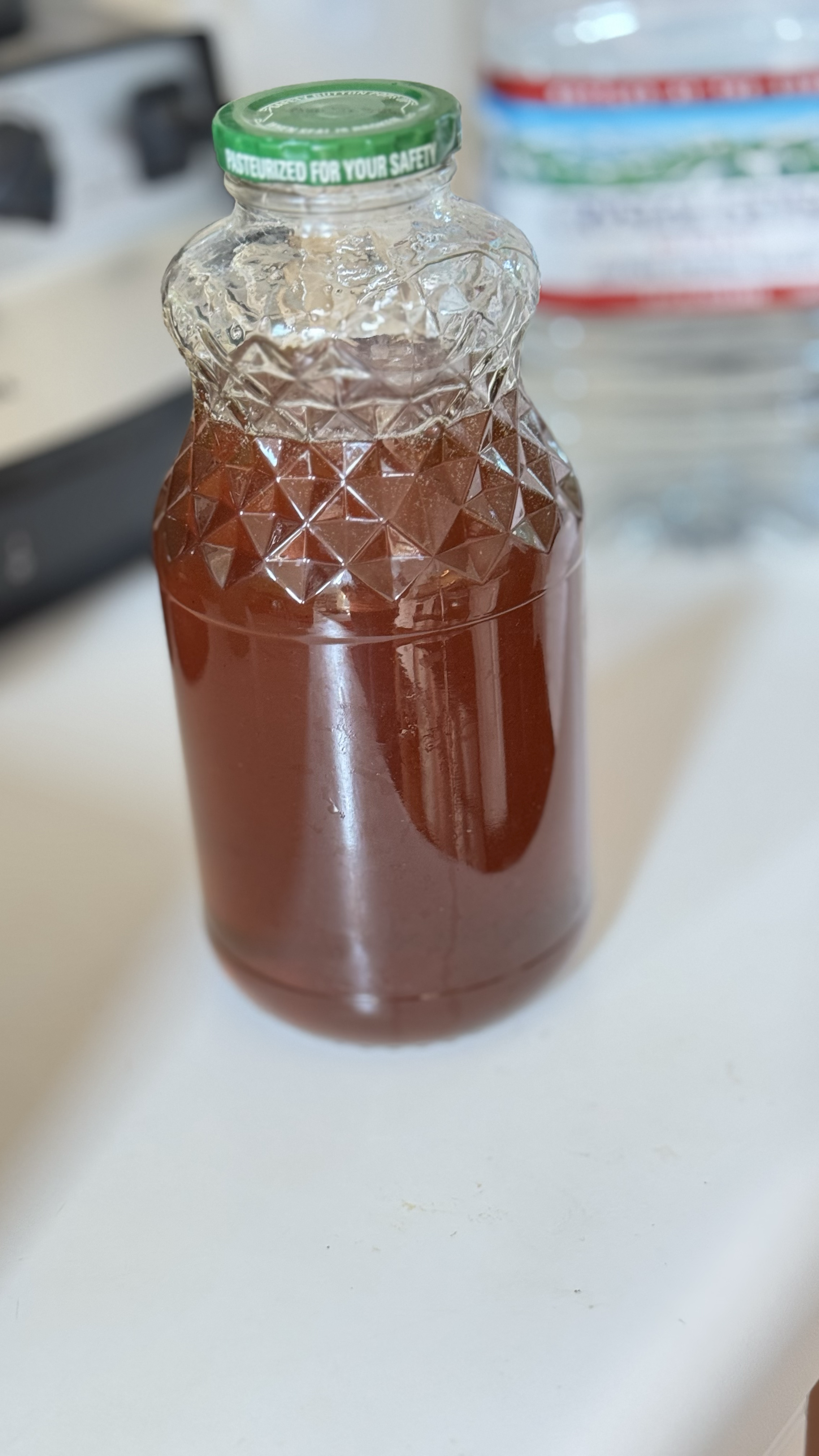 Clear glass syrup bottle filled with brown liquid on a white surface, with a green cap that says 'Pasteurized for Your Safety'.
