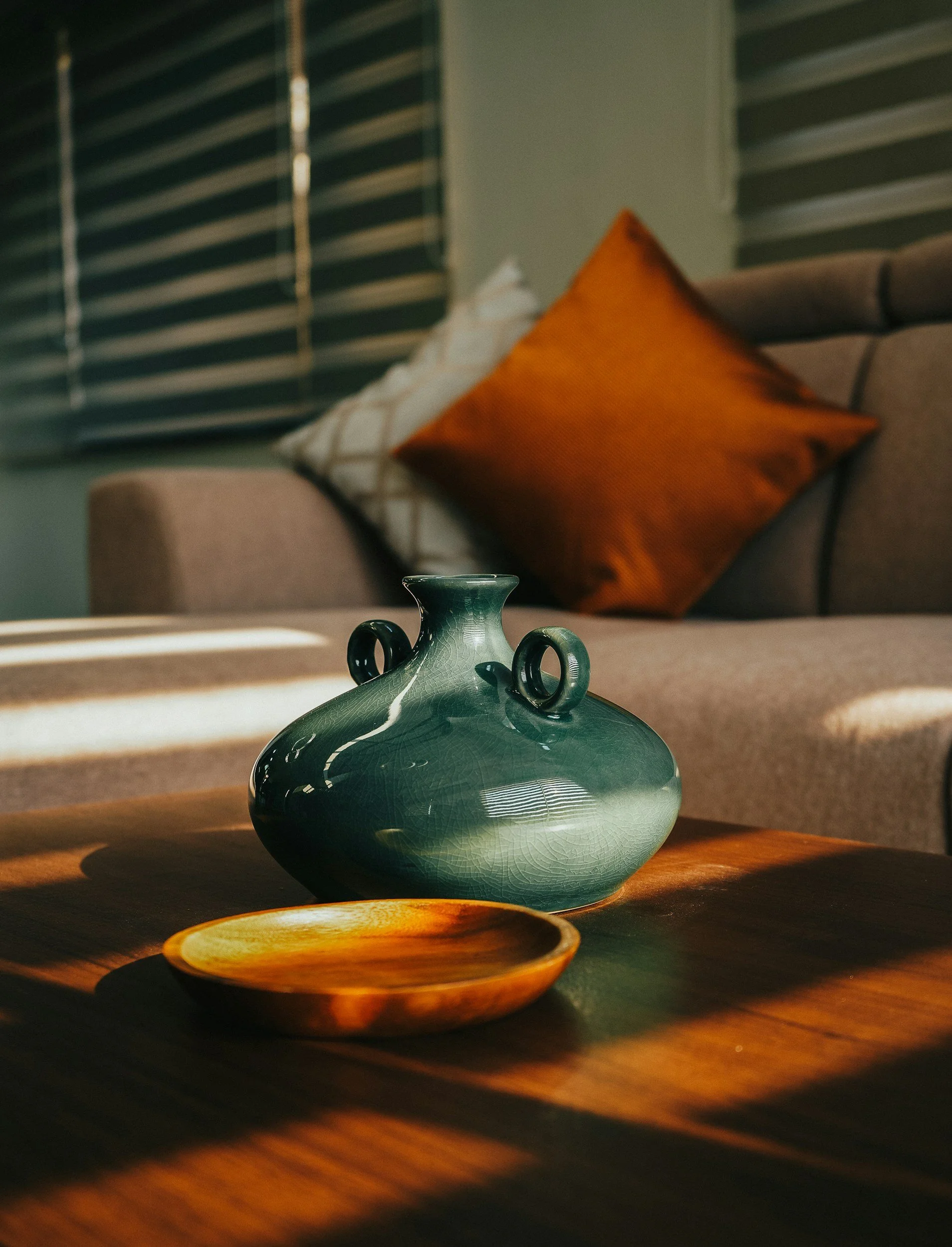 Decorative ceramic vase on a wooden table with a small wooden dish in front, inside a living room with a beige sofa and brown and white pillows, sunlight casting shadows.