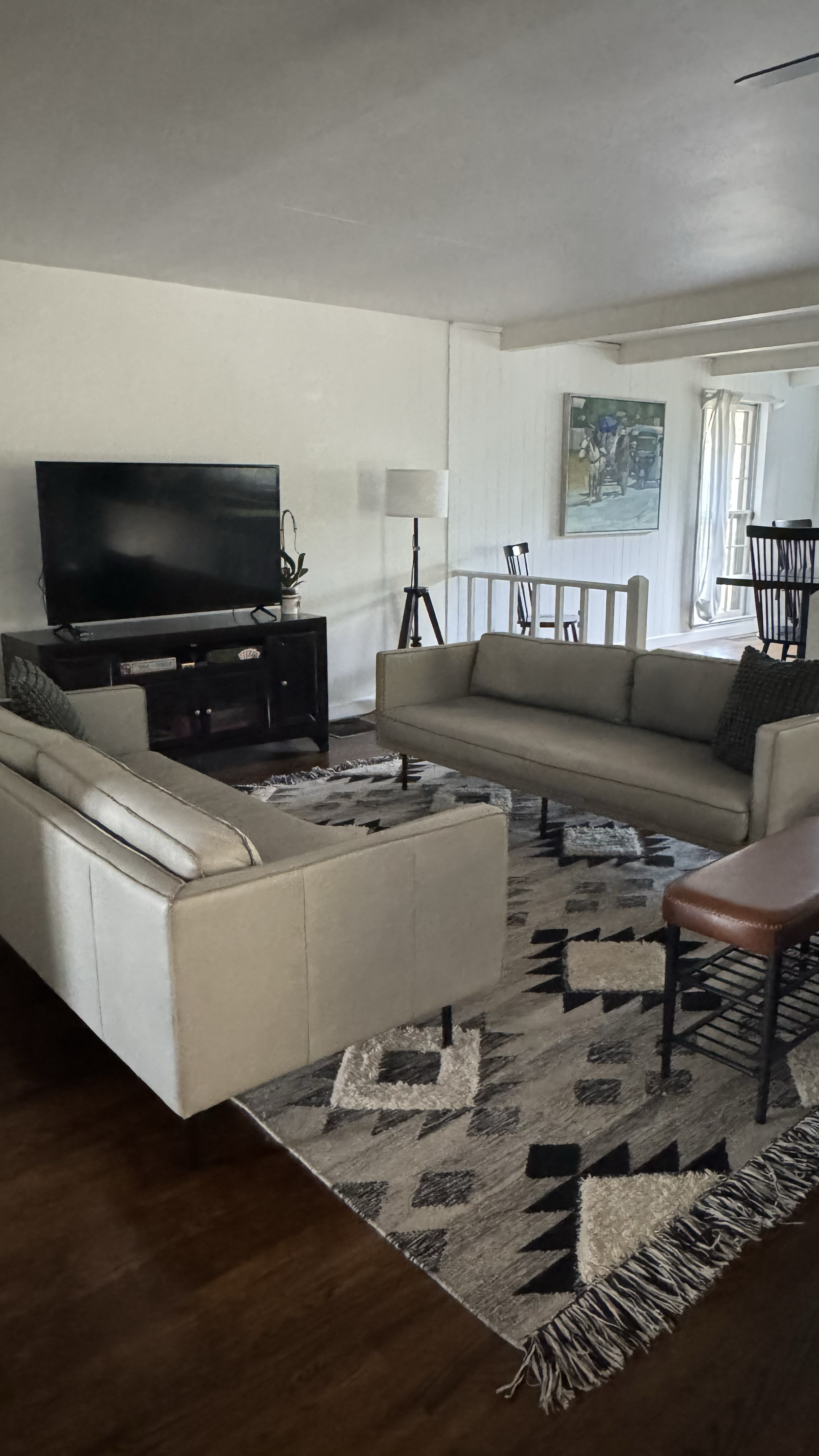 Living room with two beige sofas, a TV on a black stand, a patterned rug, a floor lamp, and a dining area with chairs near a window.