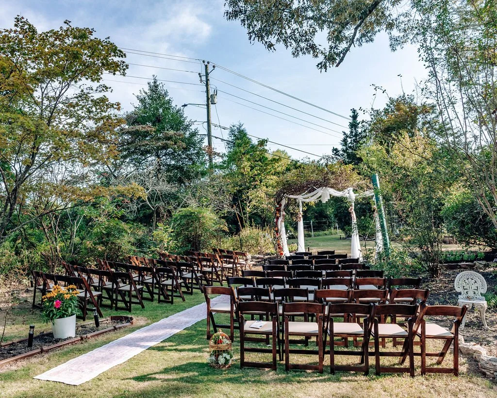 Outdoor wedding ceremony setup with rows of wooden chairs, a white aisle runner, and a decorated arch in a lush garden area surrounded by trees and greenery.