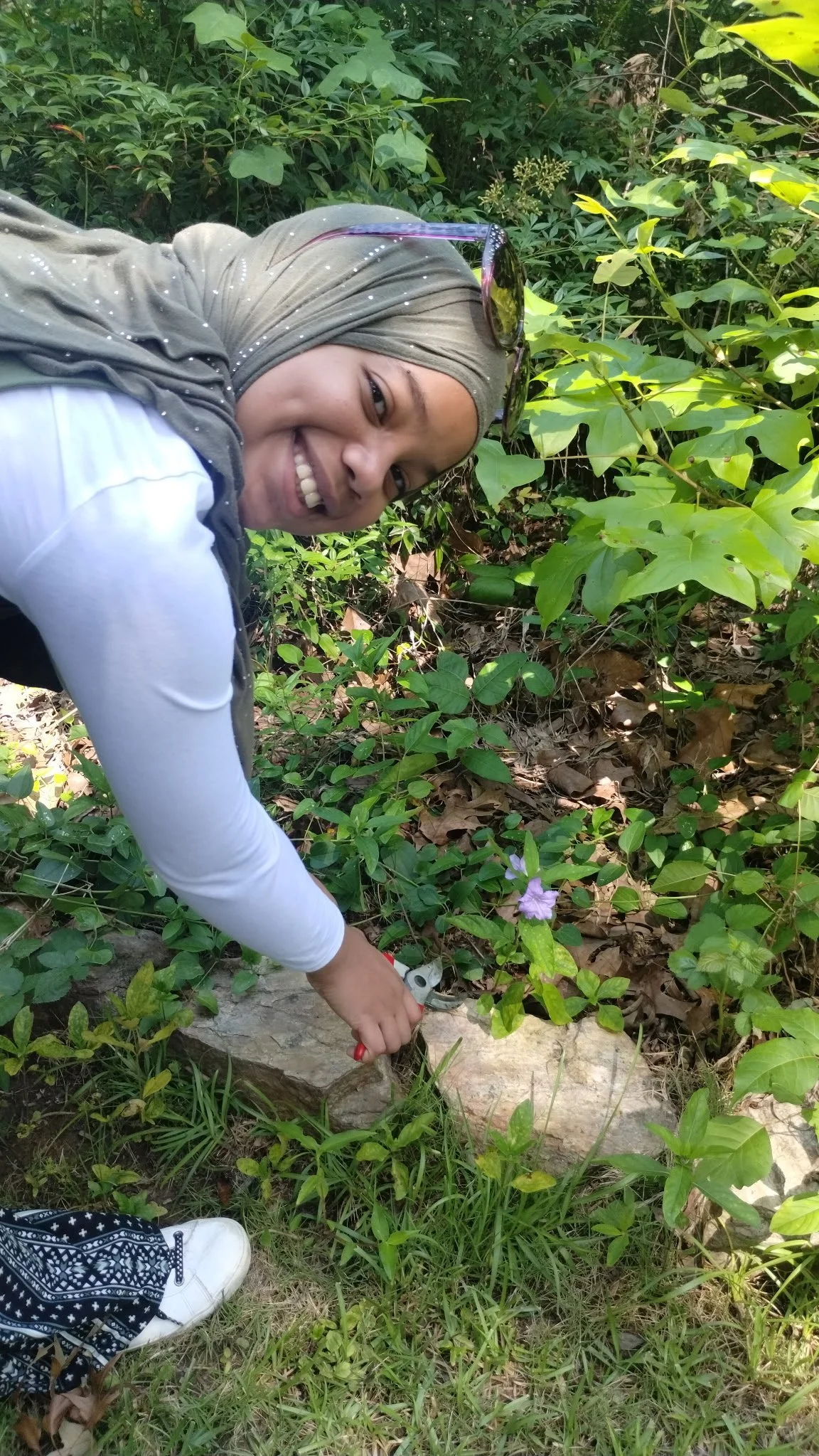 A smiling woman wearing a headscarf, sunglasses, and a white long-sleeve shirt, crouching outdoors among green foliage, holding a small gardening tool near a purple flower.