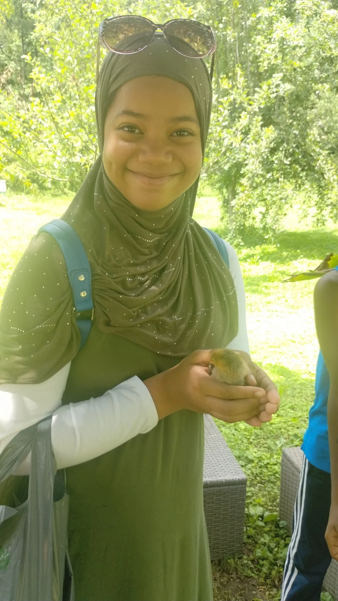 A woman wearing a hijab and sunglasses on her head, smiling and holding a small, yellow and brown duckling in her hands outdoors with trees and greenery in the background.
