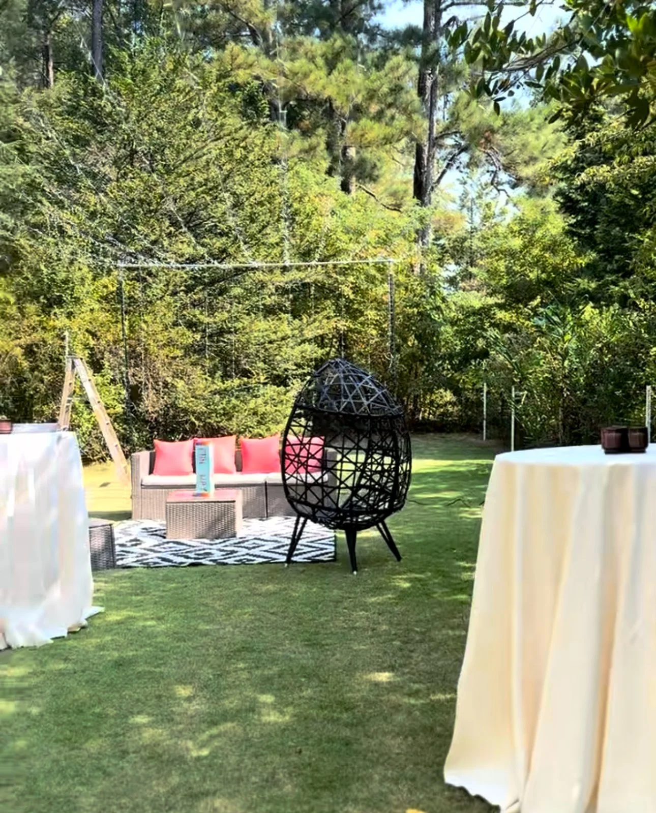 An outdoor backyard scene with a black hanging egg-shaped chair, a beige woven sofa with pink cushions, a small table, and white-draped tables set up on a grassy area. The background features tall trees and greenery.