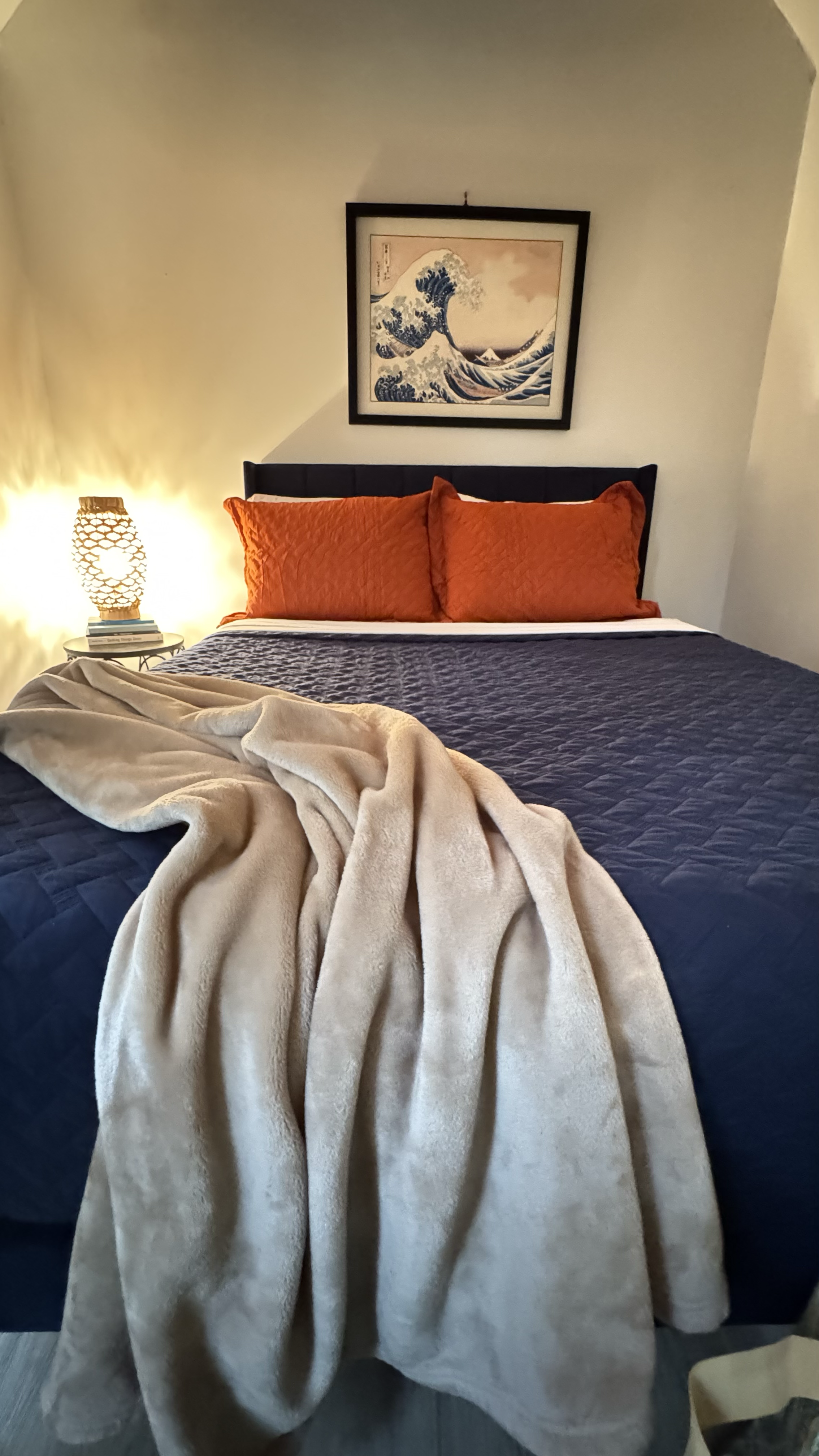 A neatly made bed with orange pillows, a navy blue quilt, and a beige blanket draped over the foot of the bed. A nightstand with a woven lamp and books is beside the bed. A framed artwork of a wave hangs above the bed.