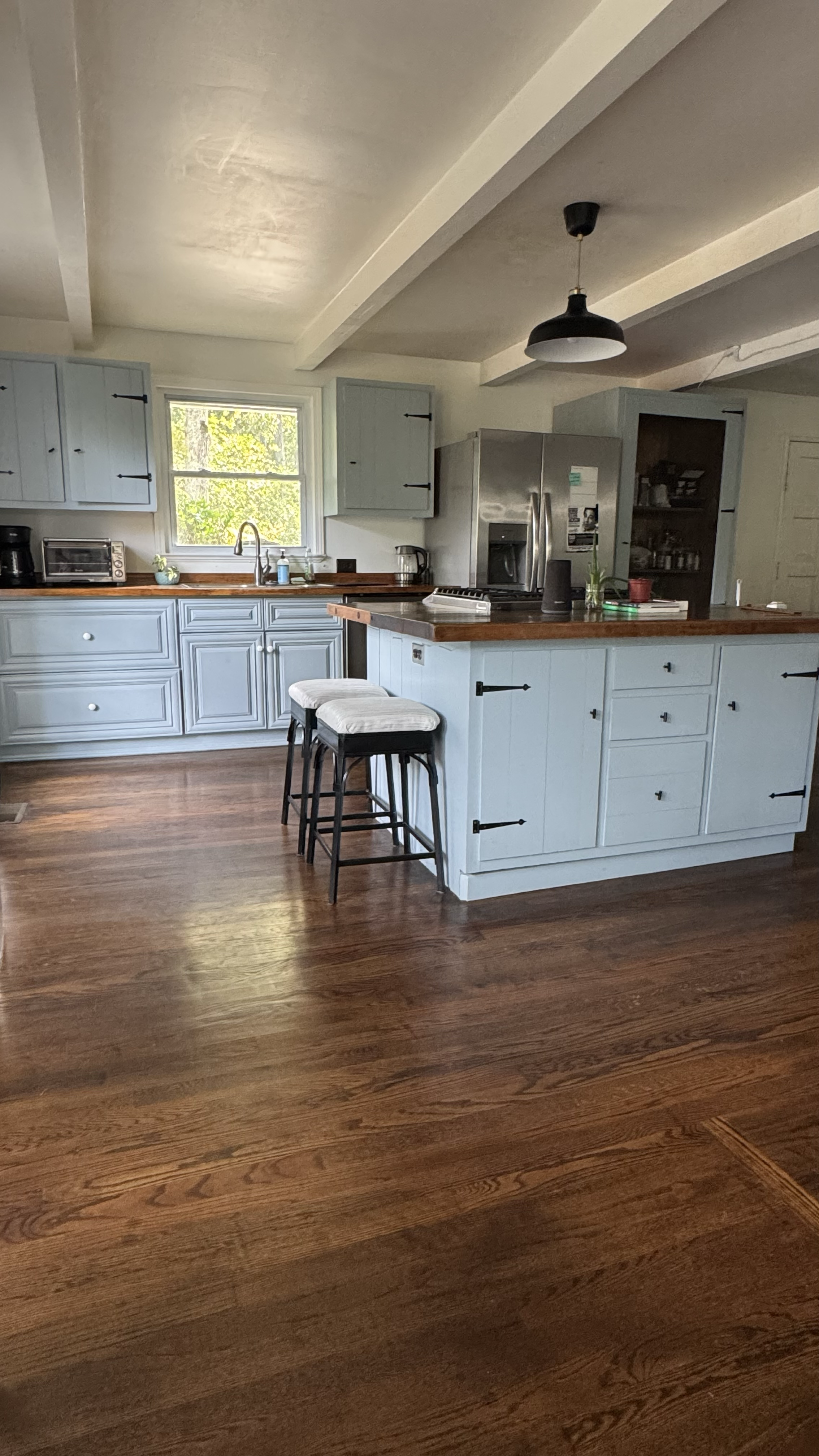 A bright kitchen with blue cabinets and a wooden countertop, hardwood flooring, a window with green trees outside, a refrigerator, and barstools at the counter.