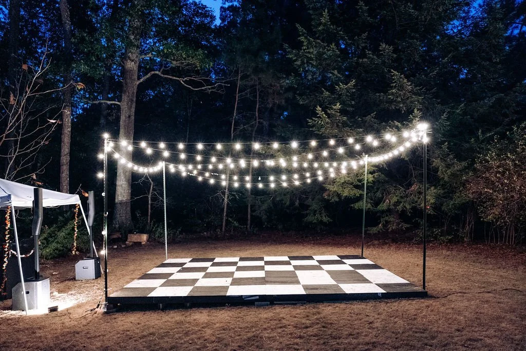 An outdoor illuminated dance floor with checkerboard pattern, string lights overhead, and a tent on the left side, set in a wooded area at night.