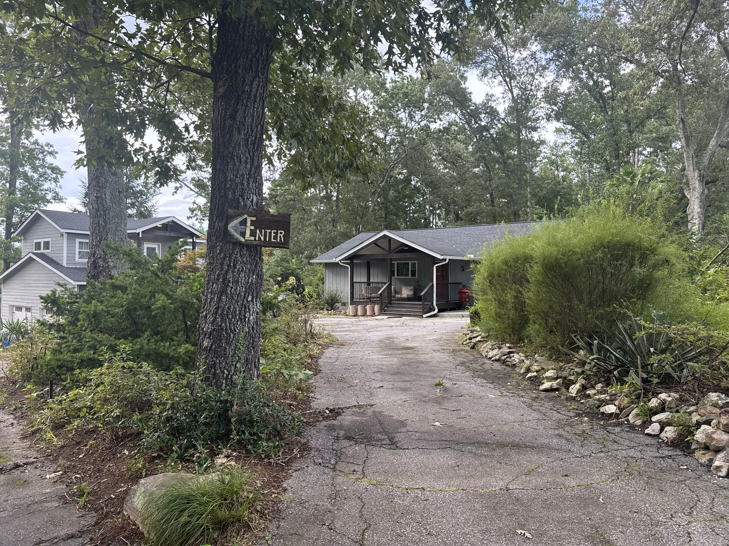 A driveway leading to a house surrounded by trees and greenery, with a wooden sign pointing to the left that says 'Enter'