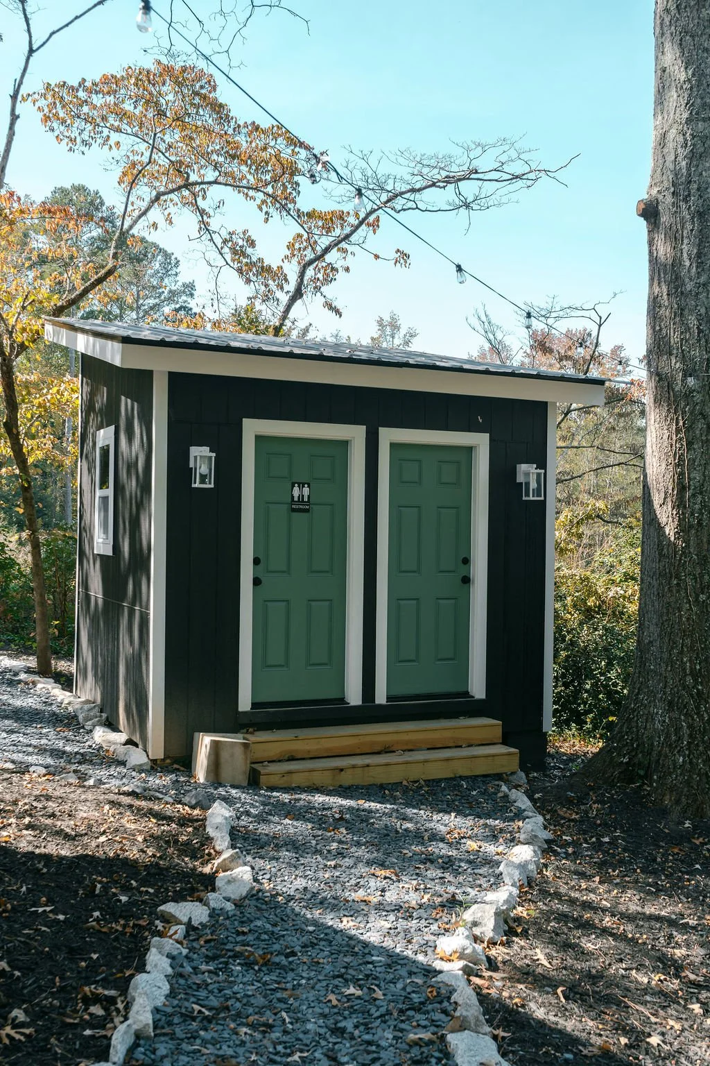 Small black outdoor bathrooms with green doors and white trim, situated in a wooded area, with a gravel path edged by white rocks leading to the entrance.