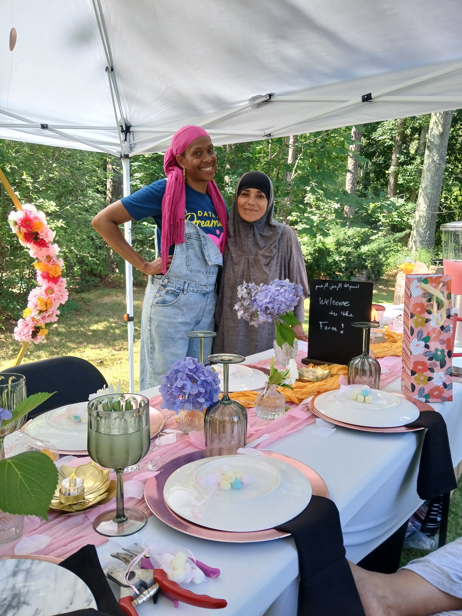 Two women standing behind a decorated outdoor table with pink, purple, and white flowers, plates, glasses, and a black chalkboard sign that says "Welcome to the Farm!" in both English and Arabic. They are smiling, under a white canopy, in a wooded ar