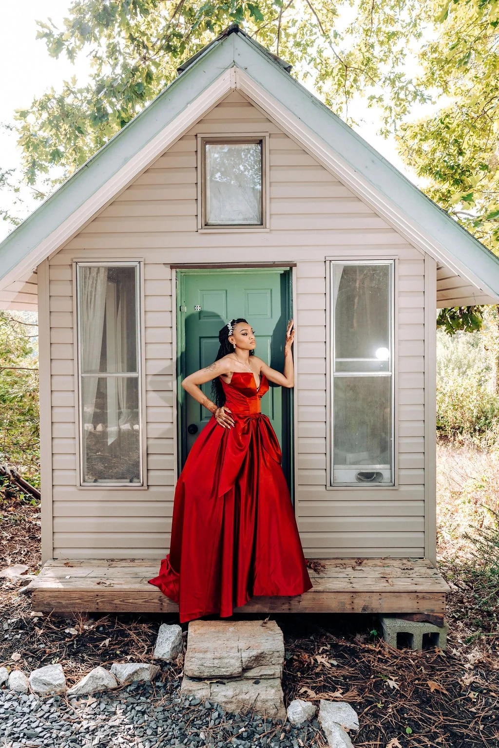 A woman in a red evening gown standing on the porch of a small beige house with a high peaked roof, surrounded by trees.