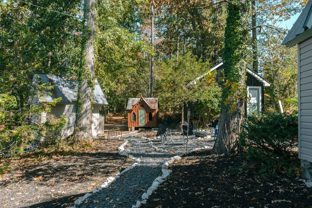 A backyard with a gravel pathway bordered by rocks, surrounded by trees, small buildings, and outdoor furniture.