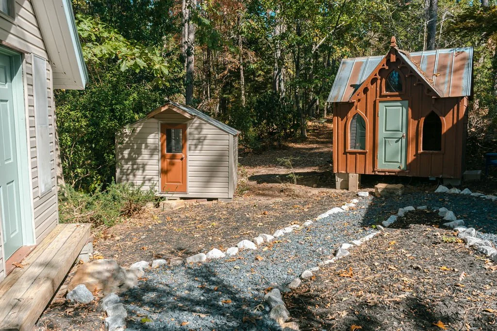 Two small decorative wooden sheds with green doors, one larger with a pointed roof, in a wooded backyard with a rocky pathway.