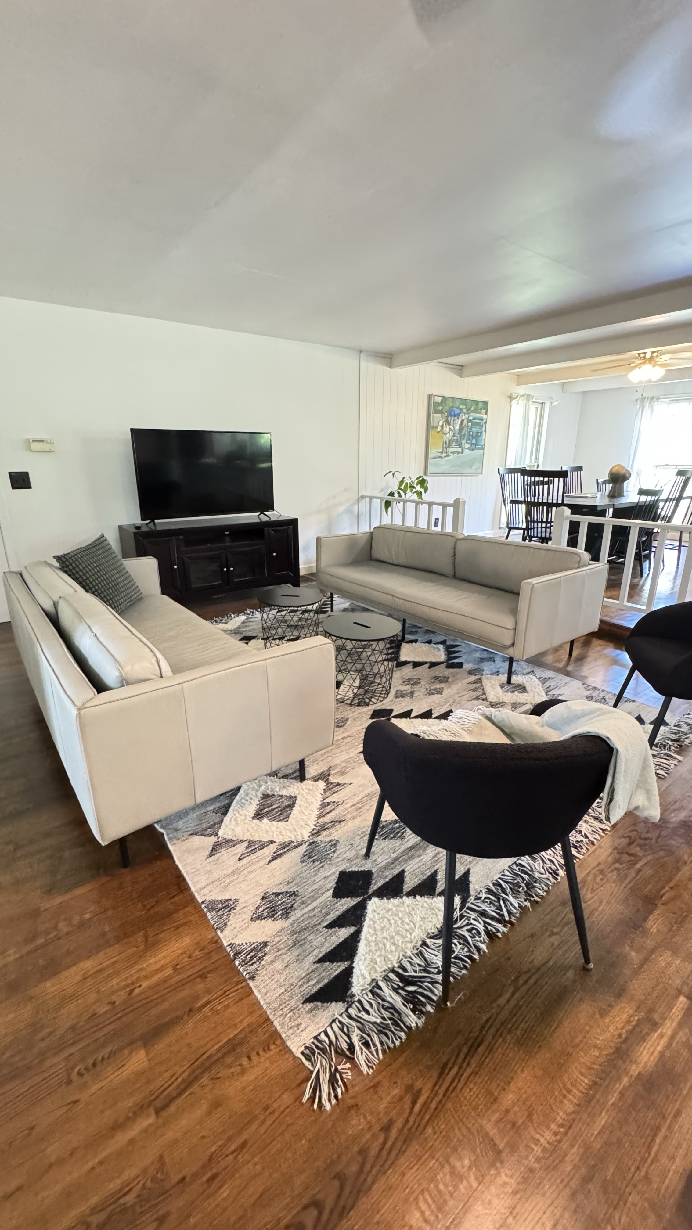 Living room with two beige sofas, black chairs, a patterned rug, a TV on a dark wood stand, and a dining area in the background.
